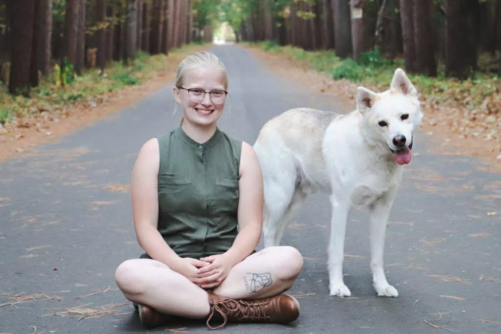 Savannah sitting on a wooded road smiling beside a calm white dog